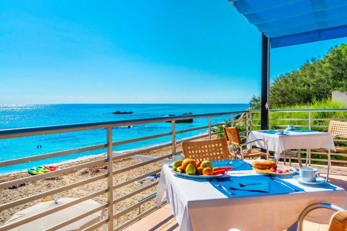 Una terraza con una mesa lista para el desayuno, con vistas a una playa de aguas azules y algunas embarcaciones.