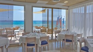 A dining area with tables set for two, large windows offering a view of the beach and ocean, and a balcony with outdoor seating. - Hotel Na Forana (Capdepera)
