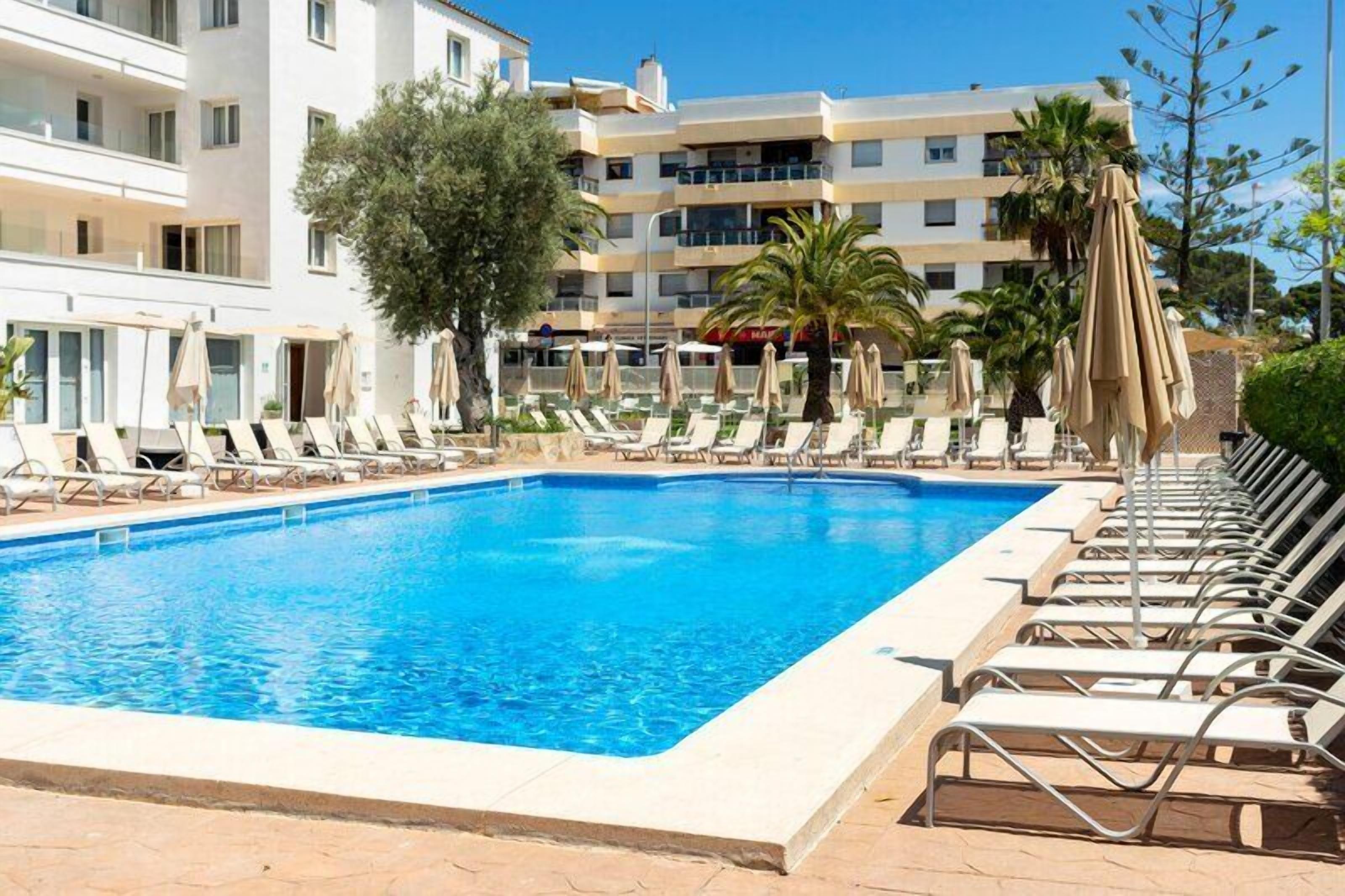 A swimming pool with lounge chairs and umbrellas, surrounded by a building with balconies and palm trees.