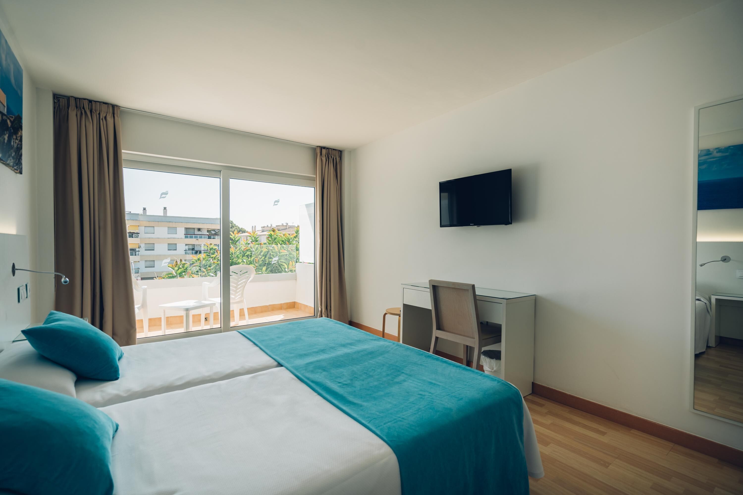 A hotel room with a bed, a desk, a chair, a TV, and a balcony with a view of buildings and greenery.