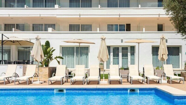 A swimming pool with lounge chairs and umbrellas, a white building with multiple windows, and a plant in the foreground. - Hotel THB Dos Playas - Adults Only (Capdepera)