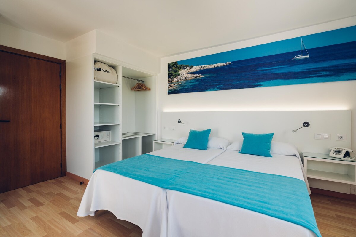 A hotel room with a bed, a wardrobe, a shelving unit with a suitcase, and a painting of a coastal scene.