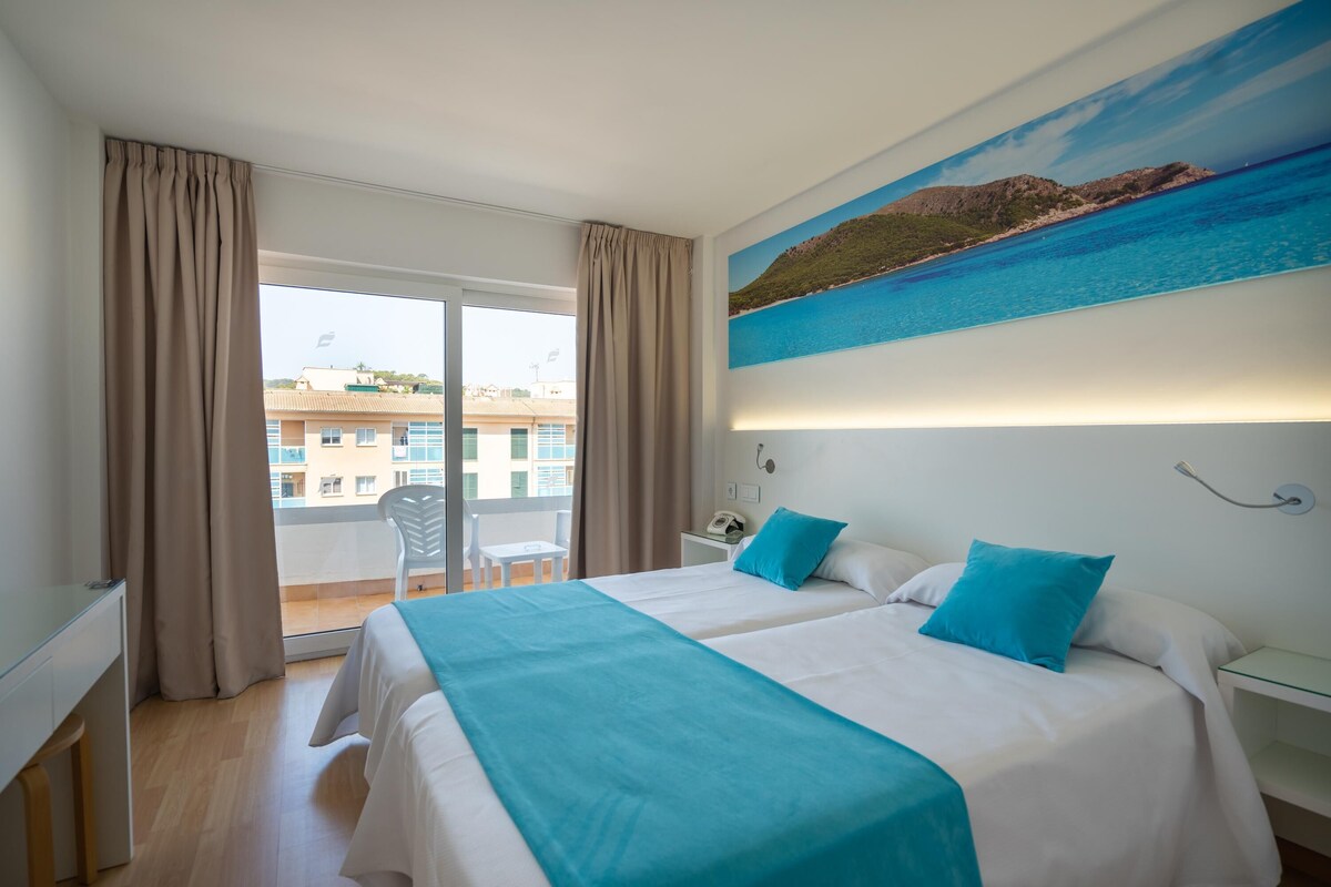 A hotel room with two beds, a balcony, a painting of a coastal landscape, and a view of buildings in the background.