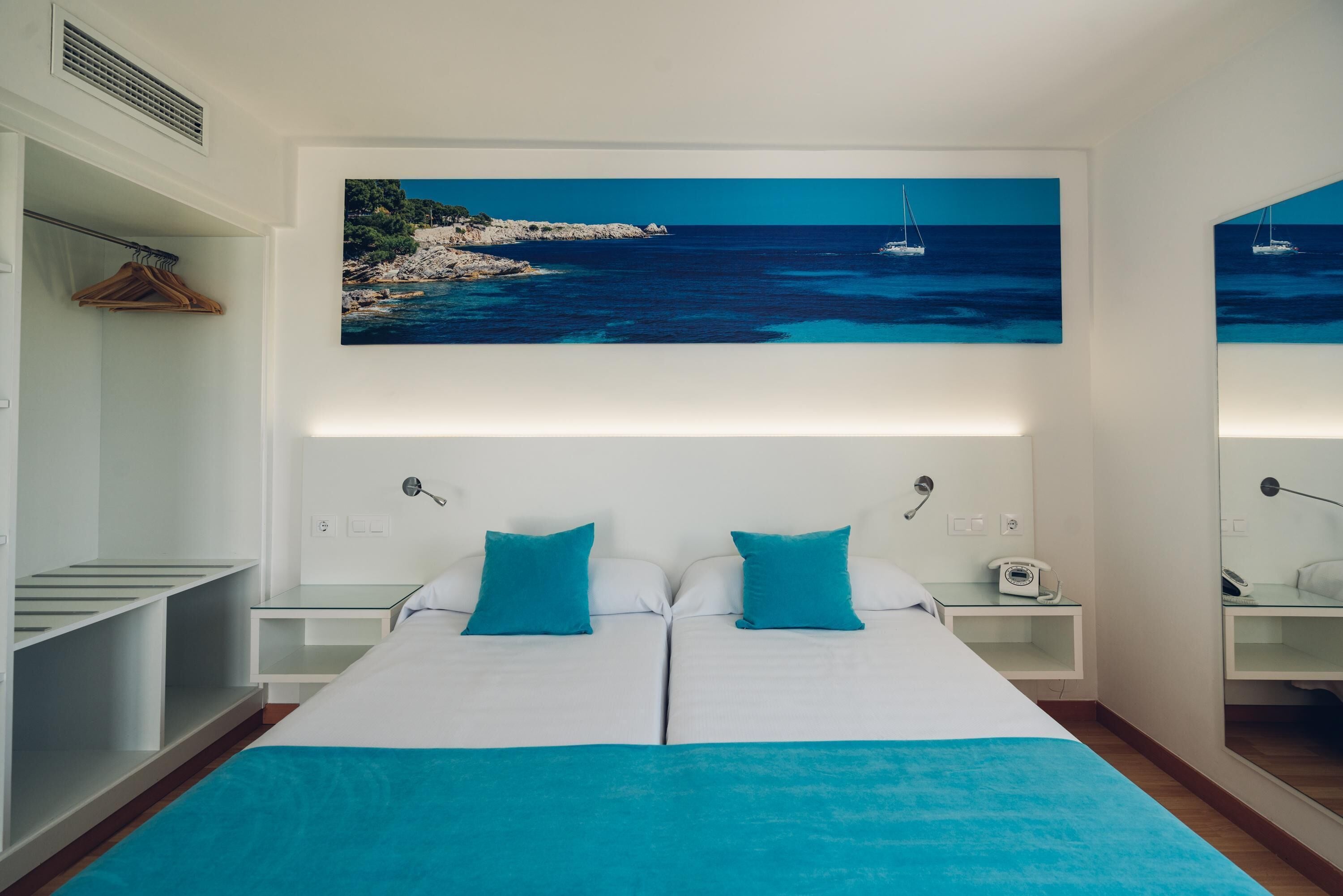Een tweepersoonsbed met twee kussens, een nachtkastje met een lamp, een kapstok en een schilderij van een kustlandschap boven het bed.