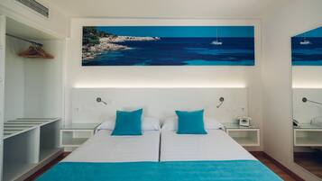 Een tweepersoonsbed met twee kussens, een nachtkastje met een lamp, een kapstok en een schilderij van een kustlandschap boven het bed.