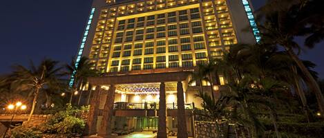 A multi-story hotel with a well-lit entrance, palm trees, and a pool area.