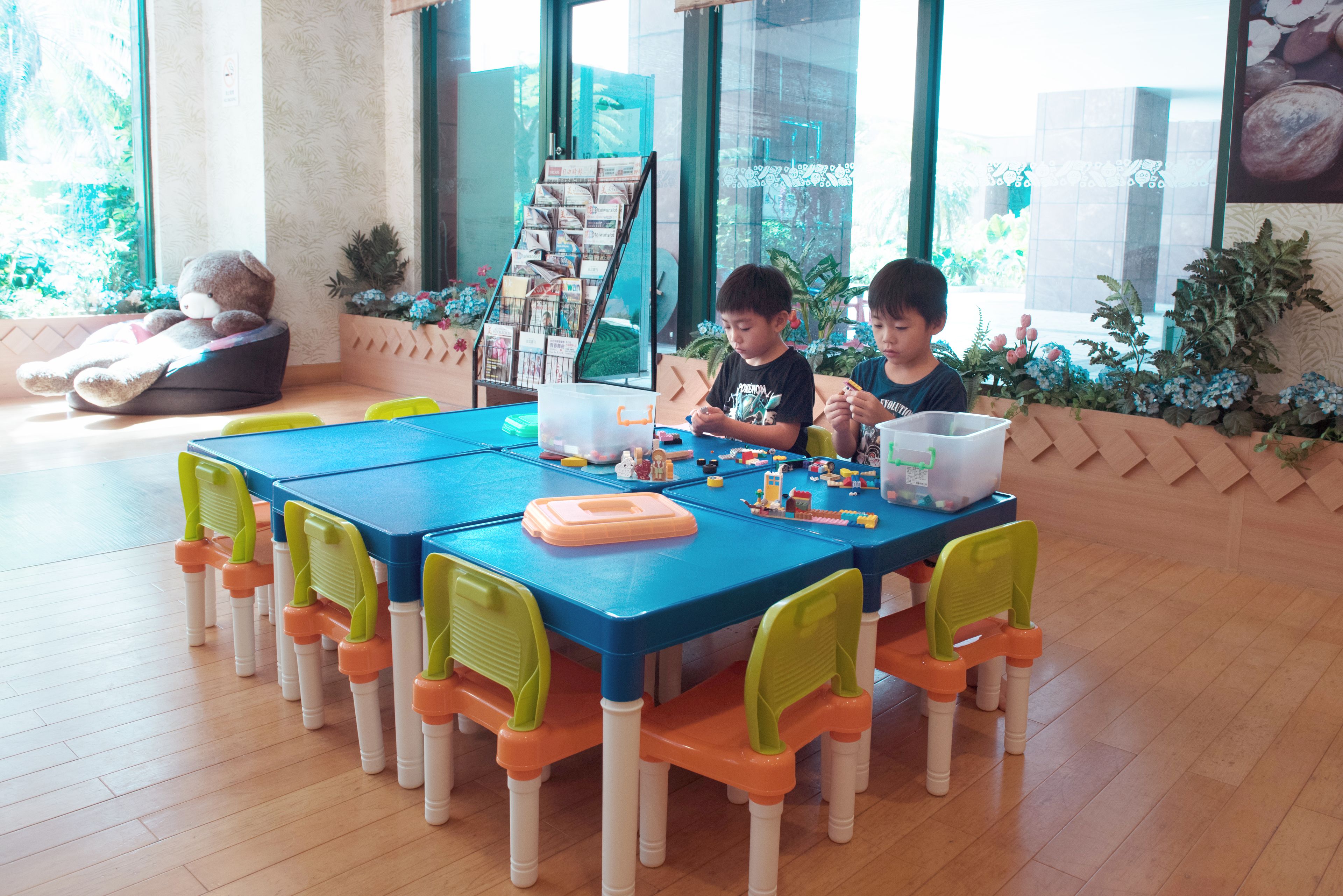Zona de juegos para niños con una mesa azul, sillas naranja y verde, y un oso de peluche sobre un cojín.