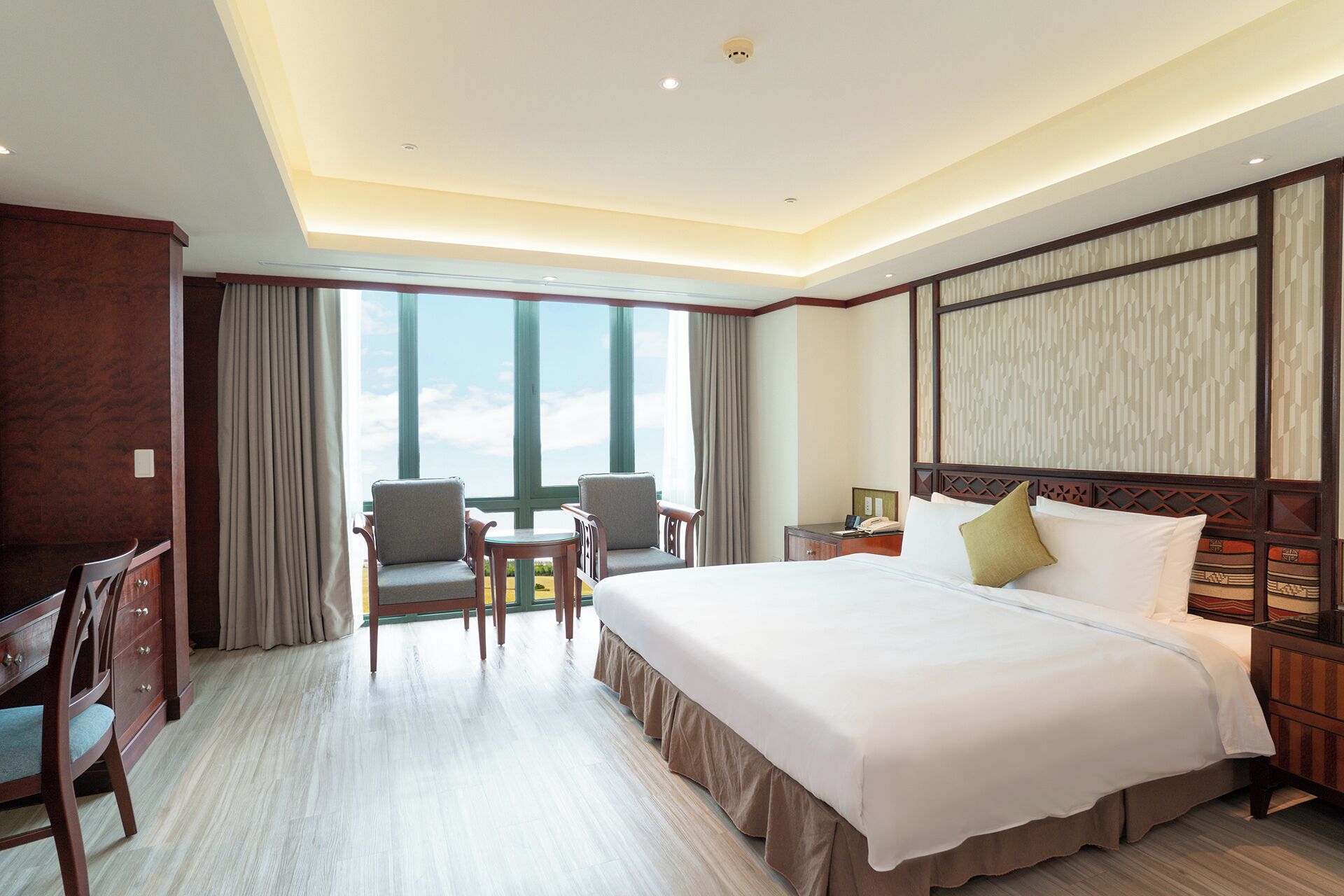 A hotel room with a large bed, a desk, two chairs, a view of the sky through large windows, and a wooden headboard.