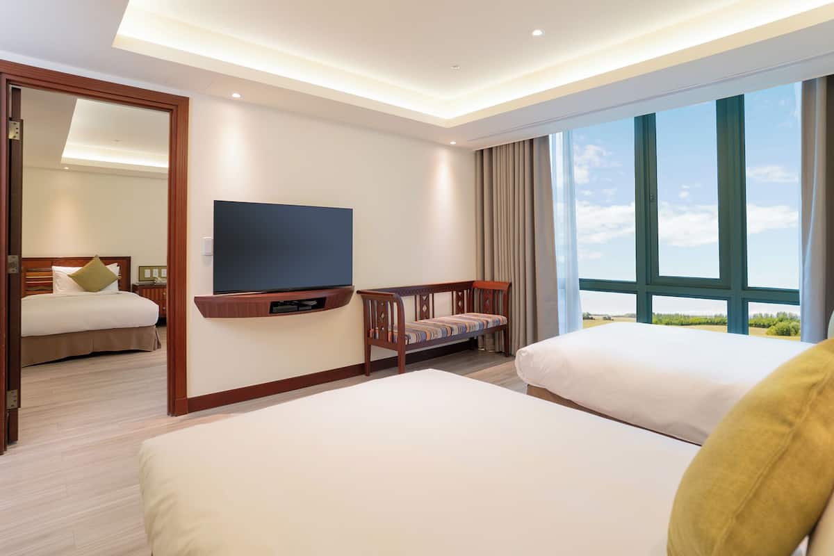 Habitació d'hotel amb un llit gran, un altre llit més petit, un televisor penjat a la paret, una taula petita i una finestra ampla que ofereix vistes a l'exterior.