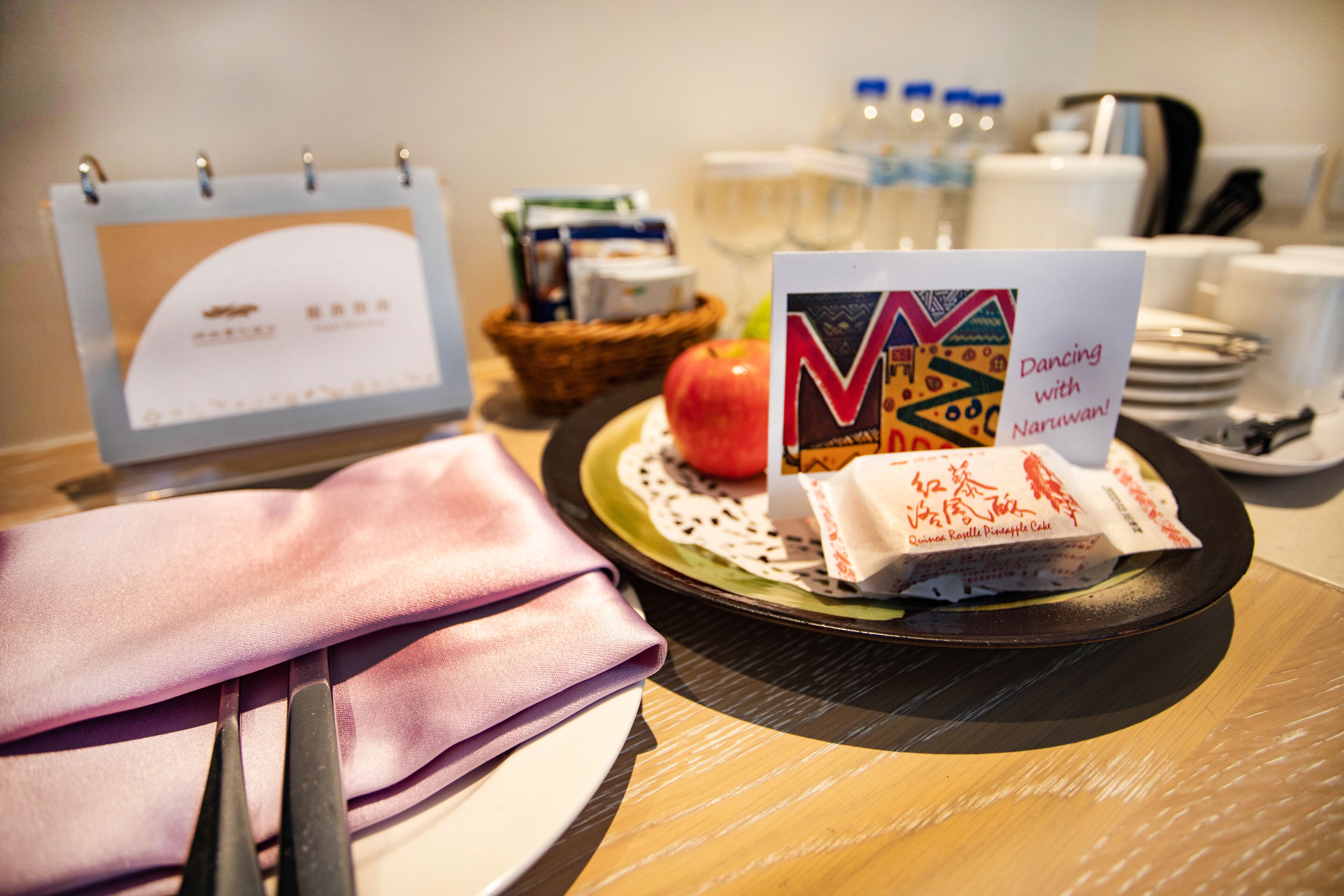 A table with a calendar, a plate with a red apple, a napkin, a pair of scissors, a plate with a card, and a basket with food items.