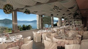 A banquet hall with round tables set for a wedding, overlooking a lake with mountains in the background. - Wellness & Beauty Hotel Conca Azzurra (Ranco)