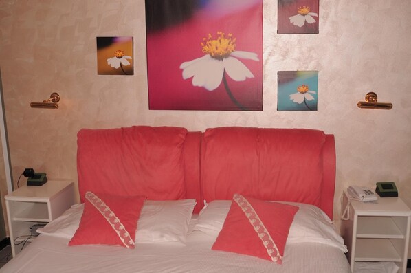 Cama doble con ropa de cama roja y almohadas blancas, dos mesitas de noche con teléfonos y un cuadro de flores en la pared.