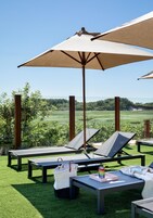 Zona junto a la piscina con sillones de descanso, una mesa con posavasos para bebidas y una sombrilla grande que da sombra.