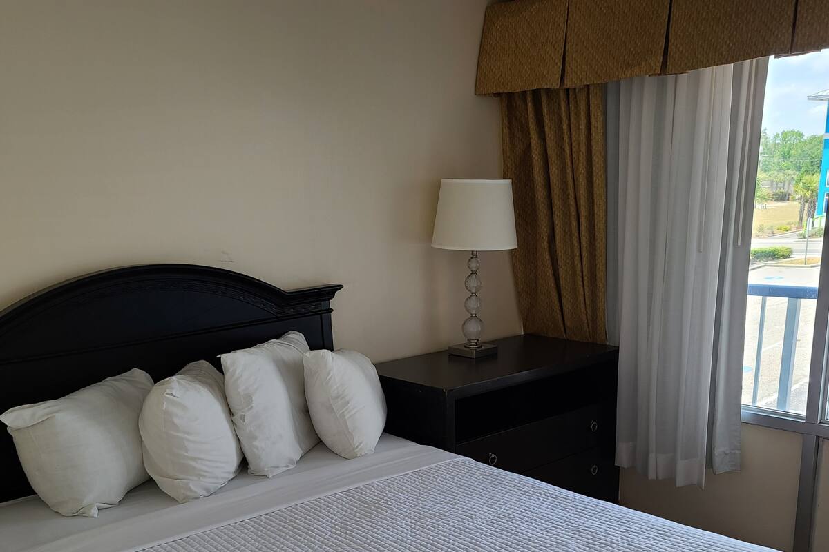 Un lit bien fait, recouvert d’une couette blanche, avec une tête de lit sombre et des oreillers blancs. On y trouve une petite table de chevet avec une lampe, et une fenêtre avec des rideaux.