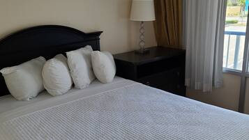 Una cama bien hecha con colcha blanca, cabecera oscura y almohadas blancas. Hay una mesita de noche con una lámpara y una ventana con cortinas.