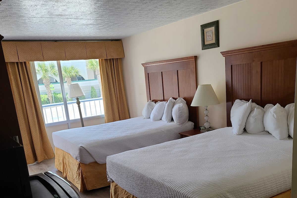 Une chambre d’hôtel avec deux lits, une tête de lit en bois, une fenêtre avec des rideaux, une lampe et un téléviseur.