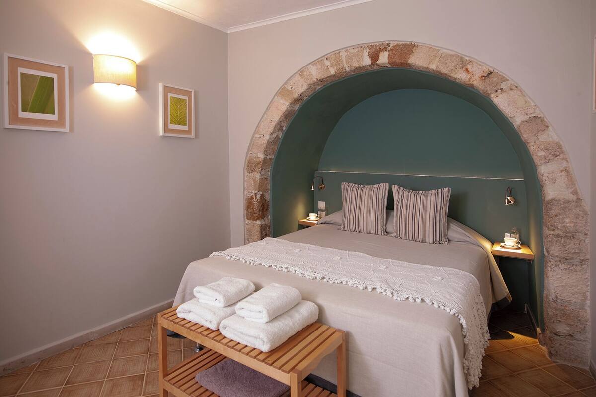 Un dormitorio con un arco de piedra, una cama con almohadas a rayas, una mesita de noche con una lámpara y un estante con toallas dobladas.