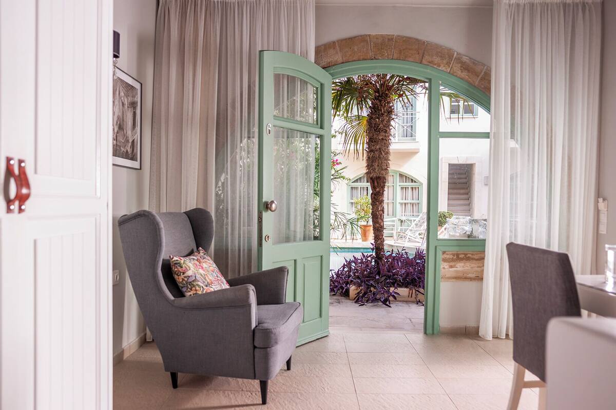 Una habitación con una puerta verde que da a una zona de piscina, un sillón gris con un cojín de colores y una mesa con sillas.