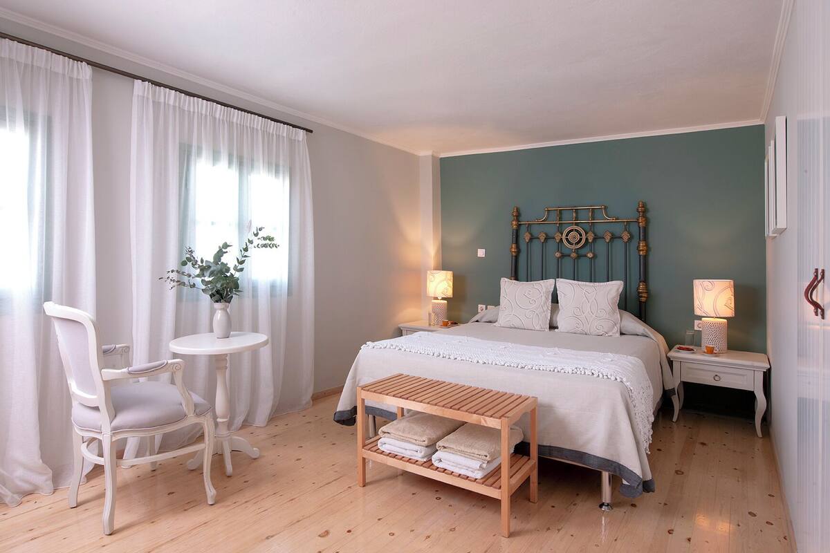 Un dormitorio con una cama, mesitas de noche, una silla, una pequeña mesa y un estante con toallas.