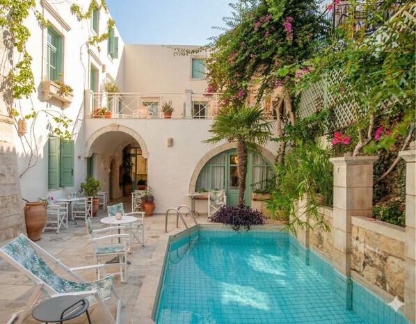 A courtyard with a swimming pool, outdoor seating, and a building with arches and balconies. - Mythos Suites Boutique Hotel (Rethymno)