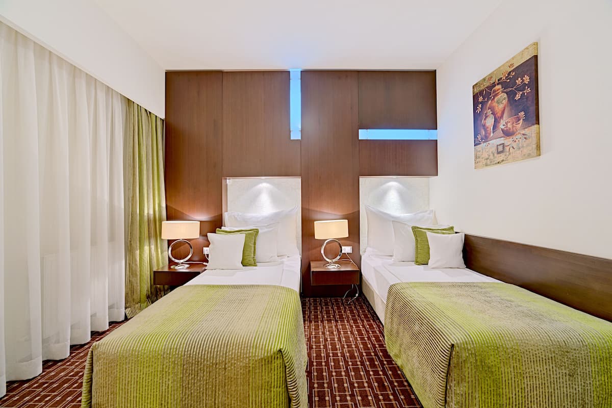 A hotel room with two beds, a large mirror, a bedside table with a lamp, and a painting on the wall.