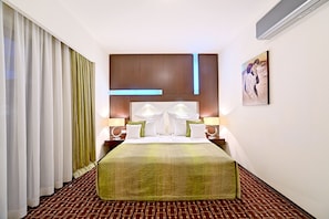 A hotel room with a large bed, a headboard with integrated lighting, a bedside table with a lamp, and a painting on the wall. - Hotel Cubix (Brasov)