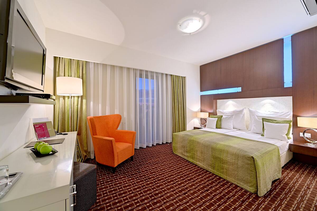 A hotel room with a bed, a desk with a computer, an orange chair, a TV, and a window with curtains.