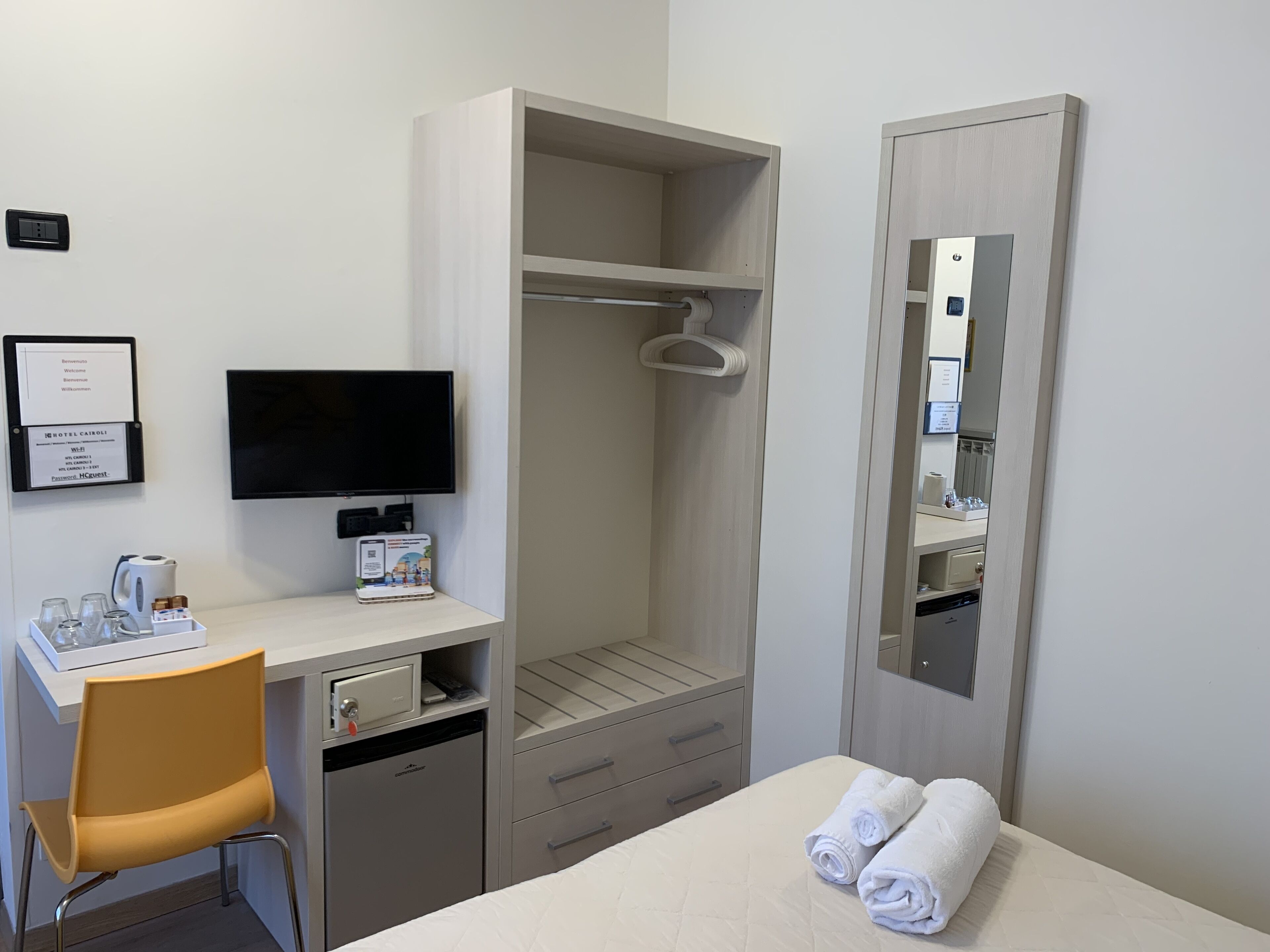 A hotel room with a desk, chair, TV, wardrobe, and a bed with towels.