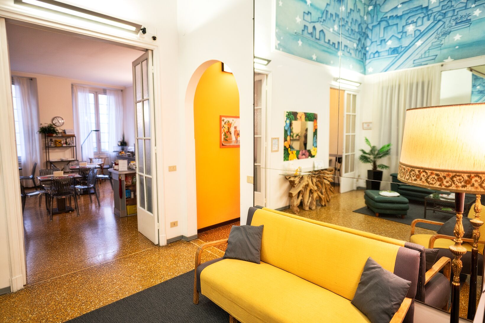 Ein hellgelbes Sofa in einem modernen Wohnzimmer mit Wandgemälde einer Stadtansicht an der Decke, einem Essbereich mit Stühlen und Tischen sowie einem Holztisch.