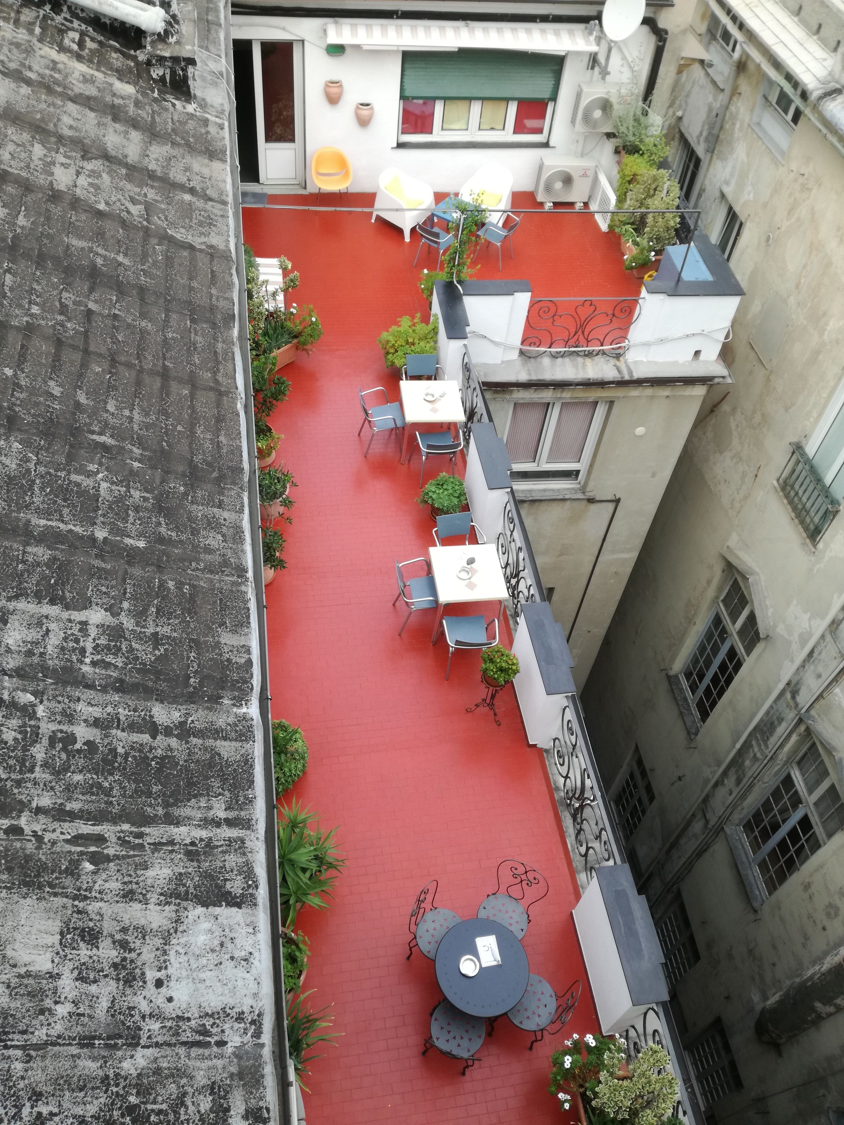 Une terrasse sur le toit avec un sol rouge, plusieurs tables et chaises, et des plantes en pot.