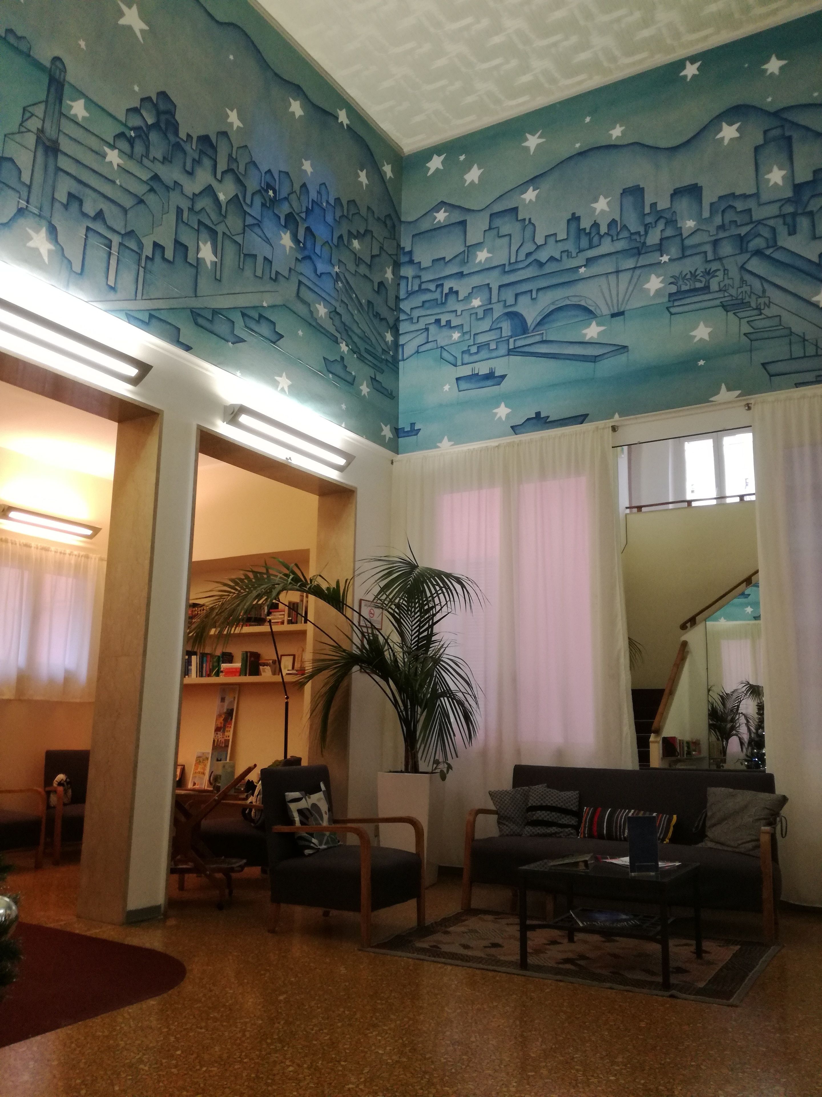 Un salón con un mural de paisaje urbano en el techo, una planta grande en maceta, un sofá y una mesa de centro.