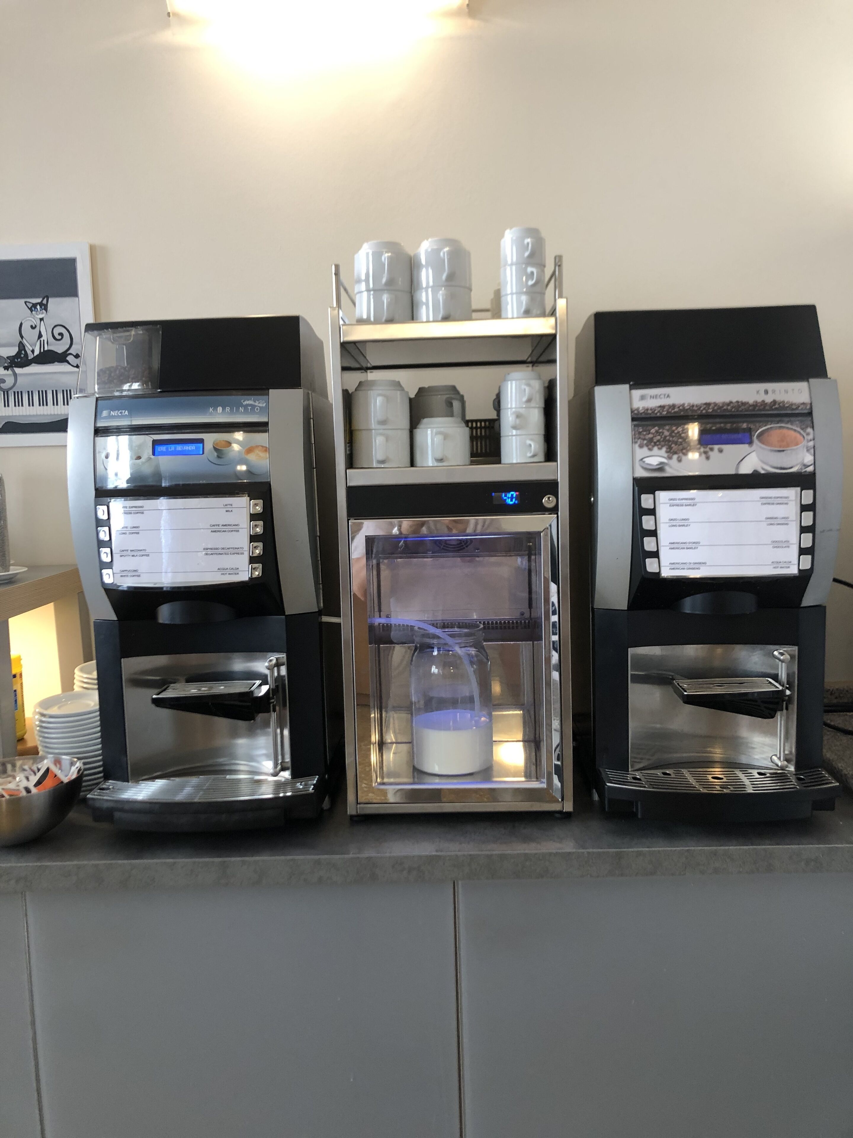 Deux machines à café, avec un appareil à faire mousser le lait placé entre les deux, une étagère au-dessus contenant des récipients à café, et un tableau encadré accroché au mur.