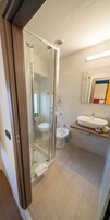 Egy modern fürőszoba üveg válaszfallal ellátott zuhanyzóval, WC-vel, bidével és egy mosdókagylóval, melynek pultja fából készült.