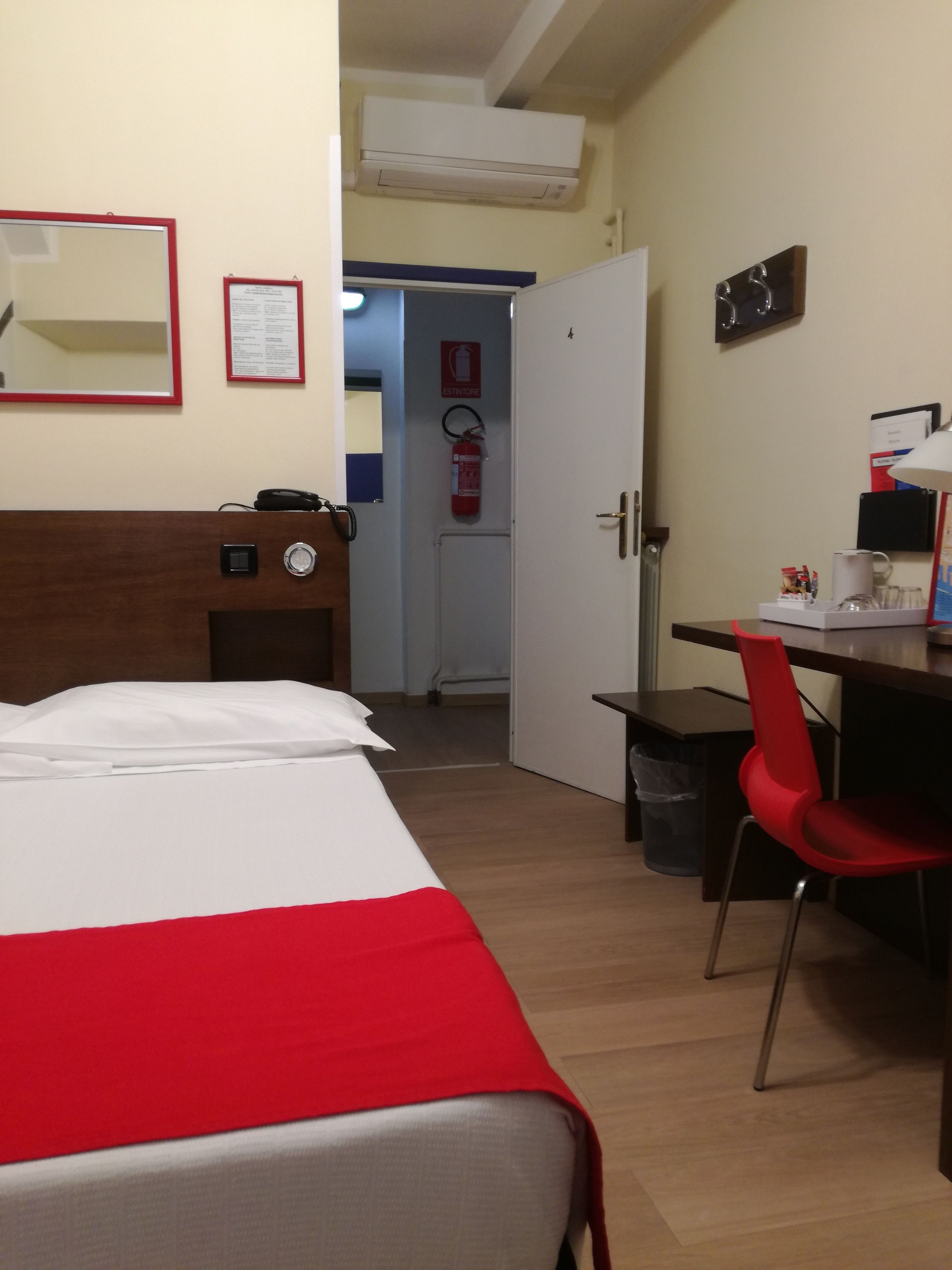 A hotel room with a bed, a desk, a chair, a mirror, a fire extinguisher, and a small trash bin.
