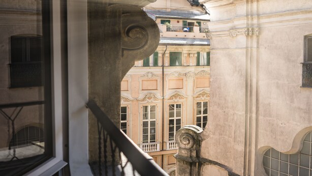 A balcony with a wrought iron railing, a stone balustrade, and a view of a building with ornate architectural details and multiple windows. - Hotel Cairoli (Genoa)