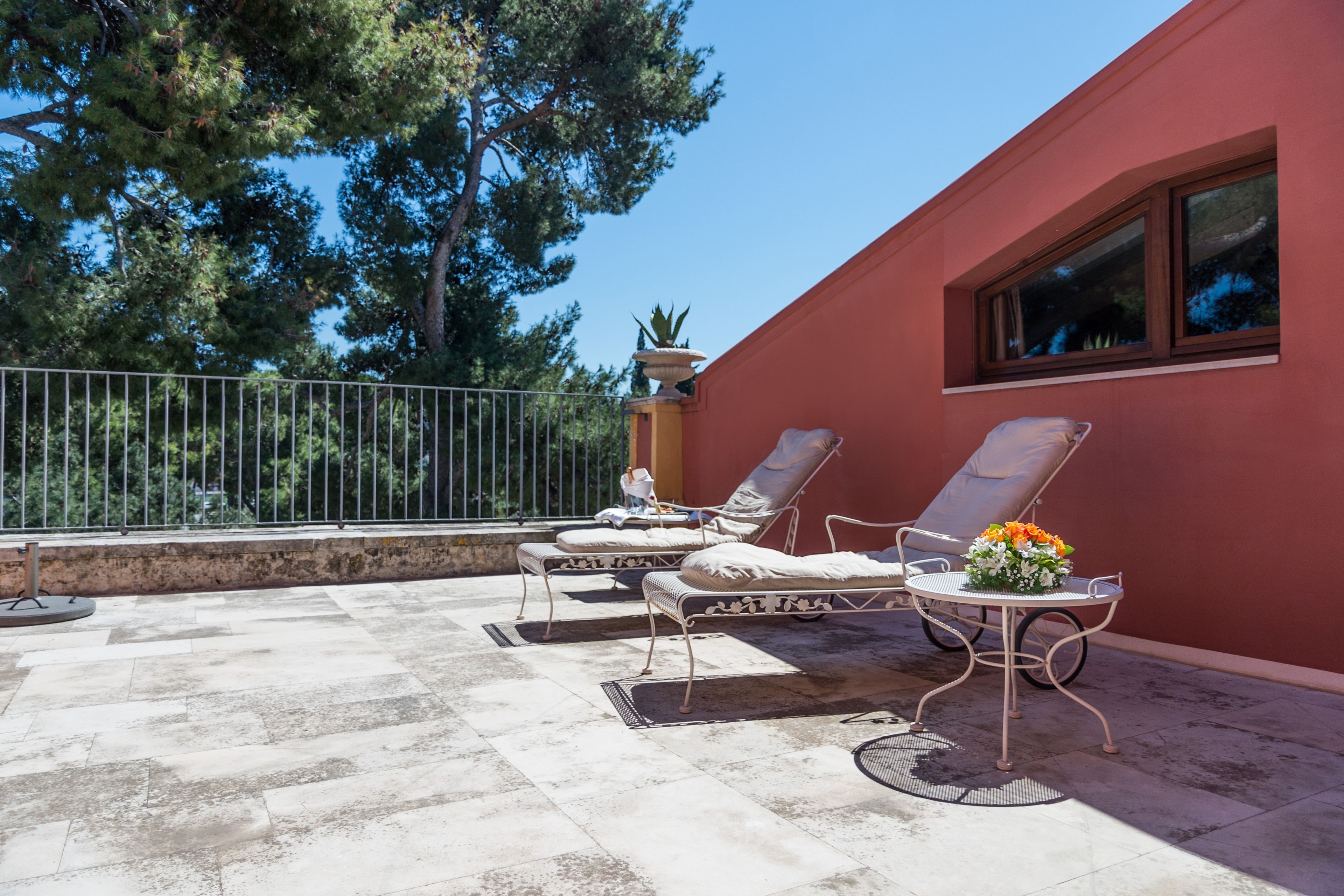 Una terraza con dos tumbonas y una mesa, rodeada por un muro rojo y una barandilla de metal.