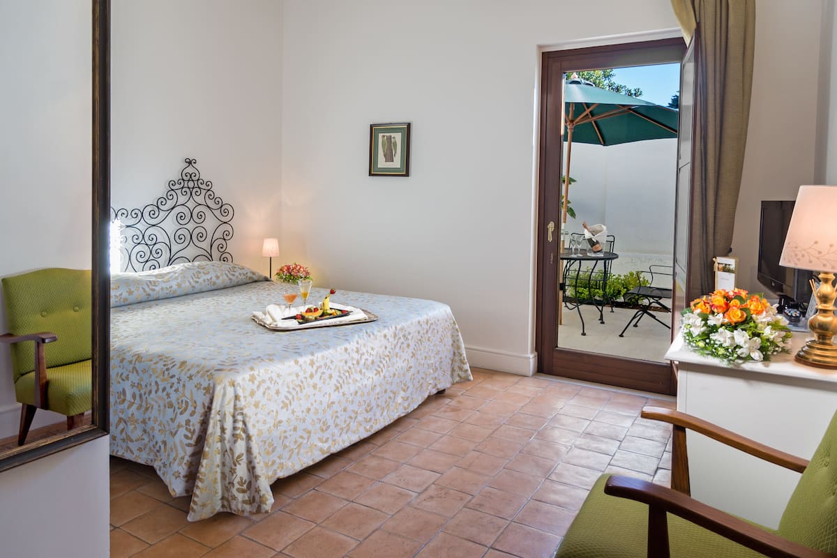 A bedroom with a bed, a bedside table with a lamp, a chair, and a view of an outdoor patio with a table and chairs.