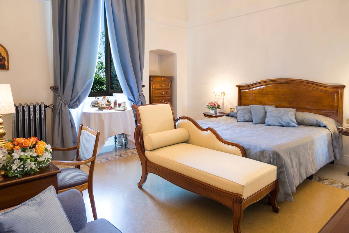 A bedroom with a large bed, a chaise lounge, a wooden dresser, and a window with curtains.