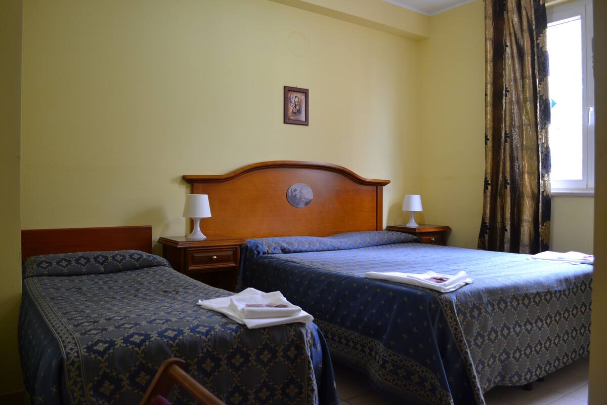 Ein Hotelzimmer mit zwei Betten, einem hölzernen Kopfteil, Nachttischen mit Lampen, einem gerahmten Bild an der Wand und einem Fenster mit Vorhängen.