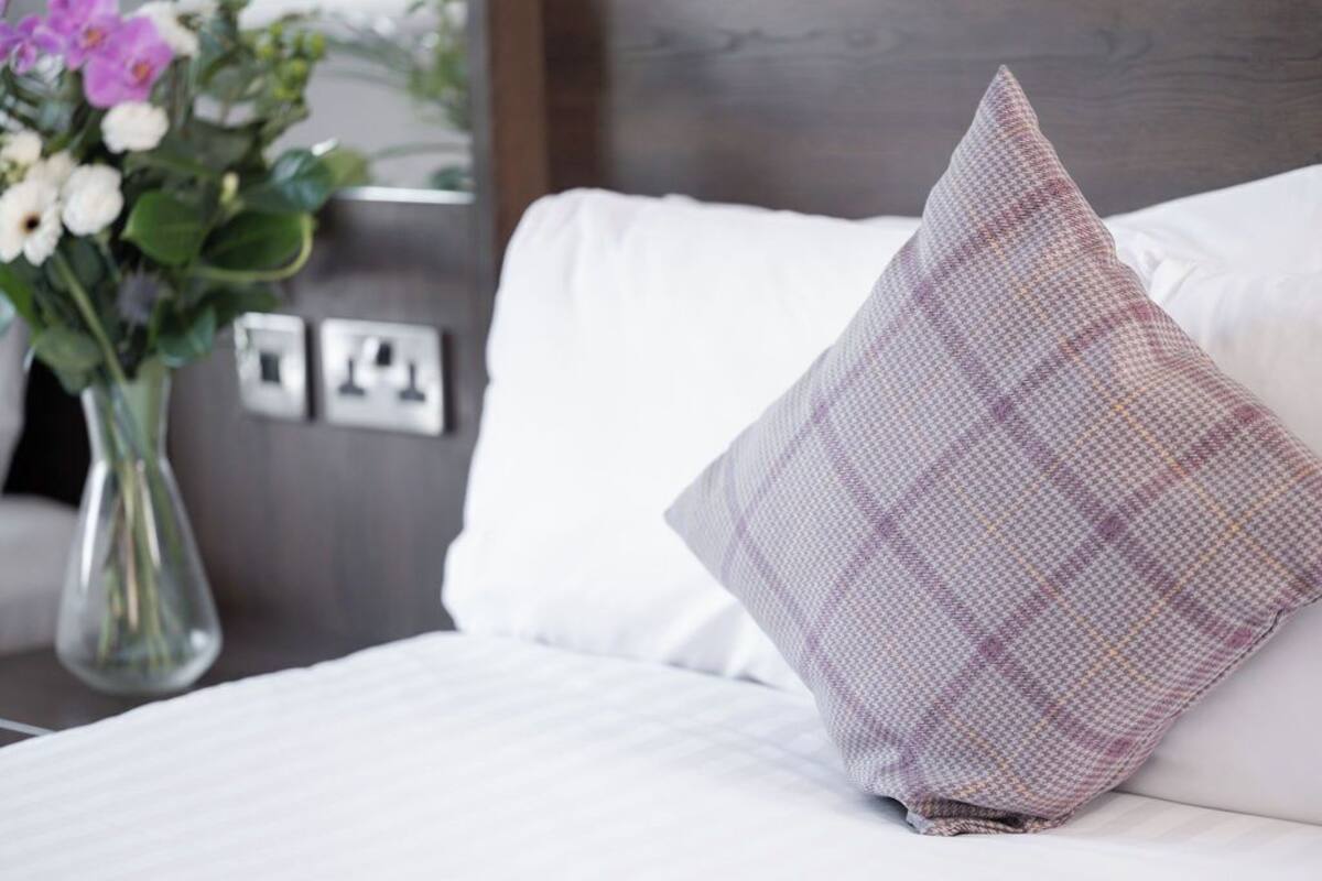 Un lit bien fait, avec du linge de lit blanc et un seul coussin à carreaux. Un vase rempli de fleurs est visible sur le côté gauche du lit.