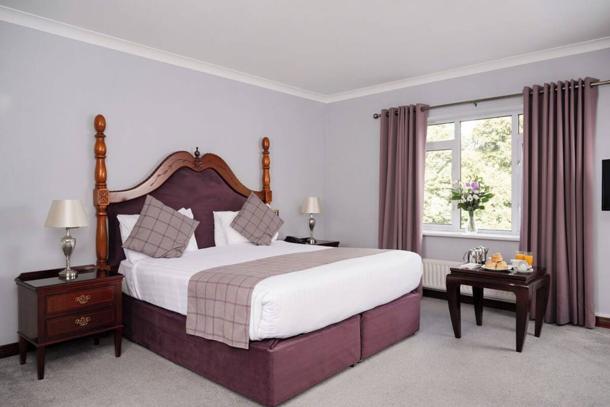 Une chambre avec un grand lit, deux tables de chevet, une fenêtre avec des rideaux et une petite table sur laquelle sont disposés des éléments pour le petit-déjeuner.