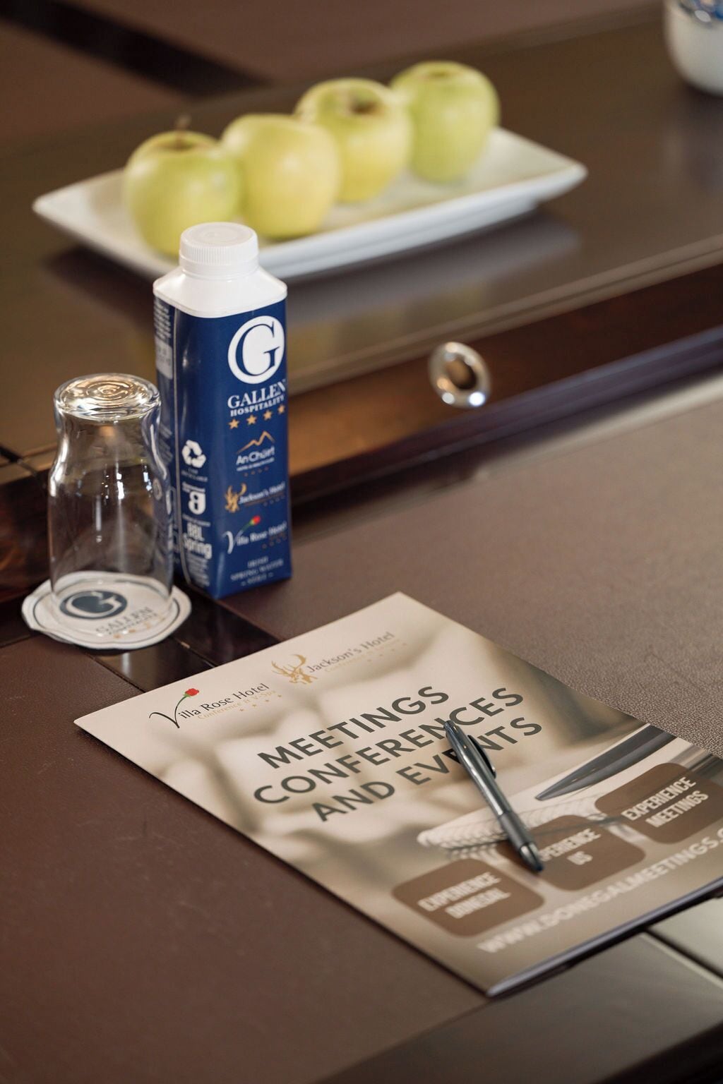 Een hotel receptiebalie met een schaal groene appels, een fles GALLEN HOSPITALITY, een glas water, en een brochure voor Villa Rose Hotel bijeenkomsten, congressen en evenementen.