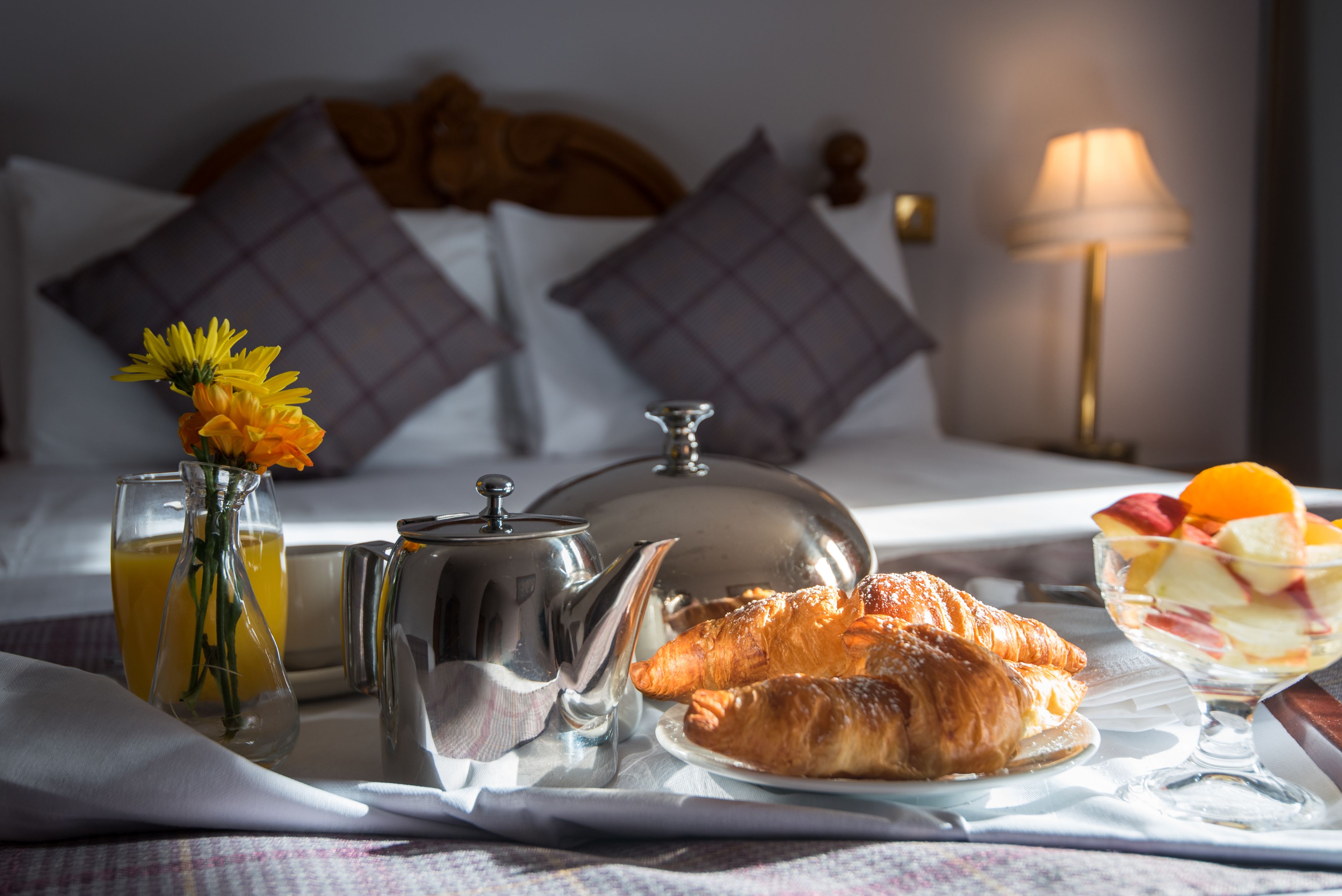 A breakfast tray with croissants, fruit, and a teapot on a bed with pillows and a lamp in a hotel room.