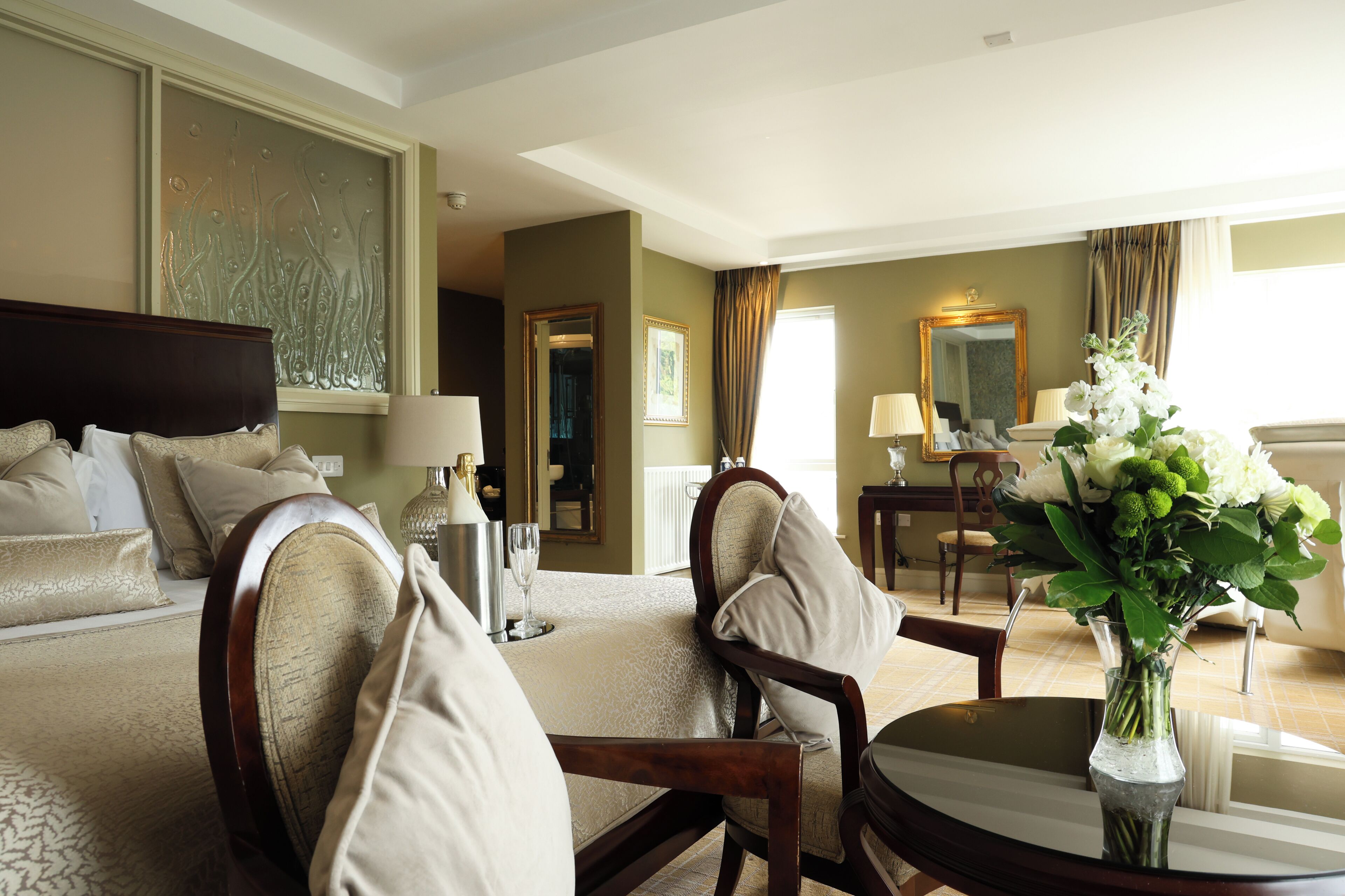 A hotel room with a bed, a sitting area with a table and chairs, a large mirror, and a window with curtains.