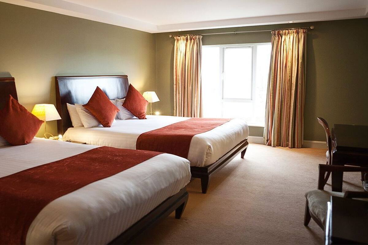 Une chambre d’hôtel avec deux lits, chacun doté d’une tête de lit en bois, de draps blancs et de coussins décoratifs rouges. Il y a une chaise et une lampe d’un côté de la pièce.