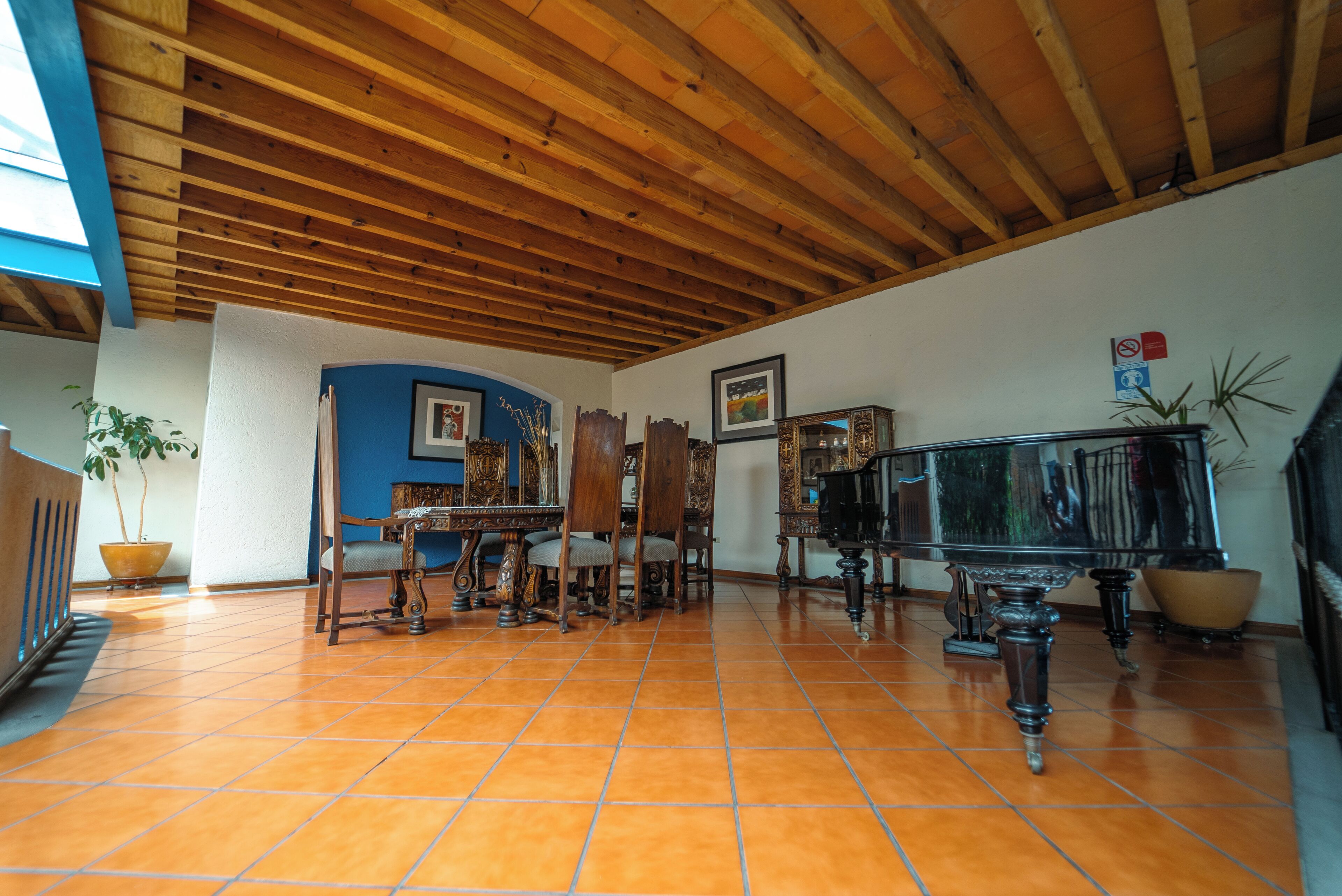 Una habitación amplia con suelo de baldosas, vigas de madera en el techo, una mesa de comedor con sillas, un piano y una planta en maceta.