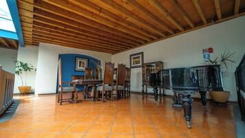 Una habitación amplia con suelo de baldosas, vigas de madera en el techo, una mesa de comedor con sillas, un piano y una planta en maceta.