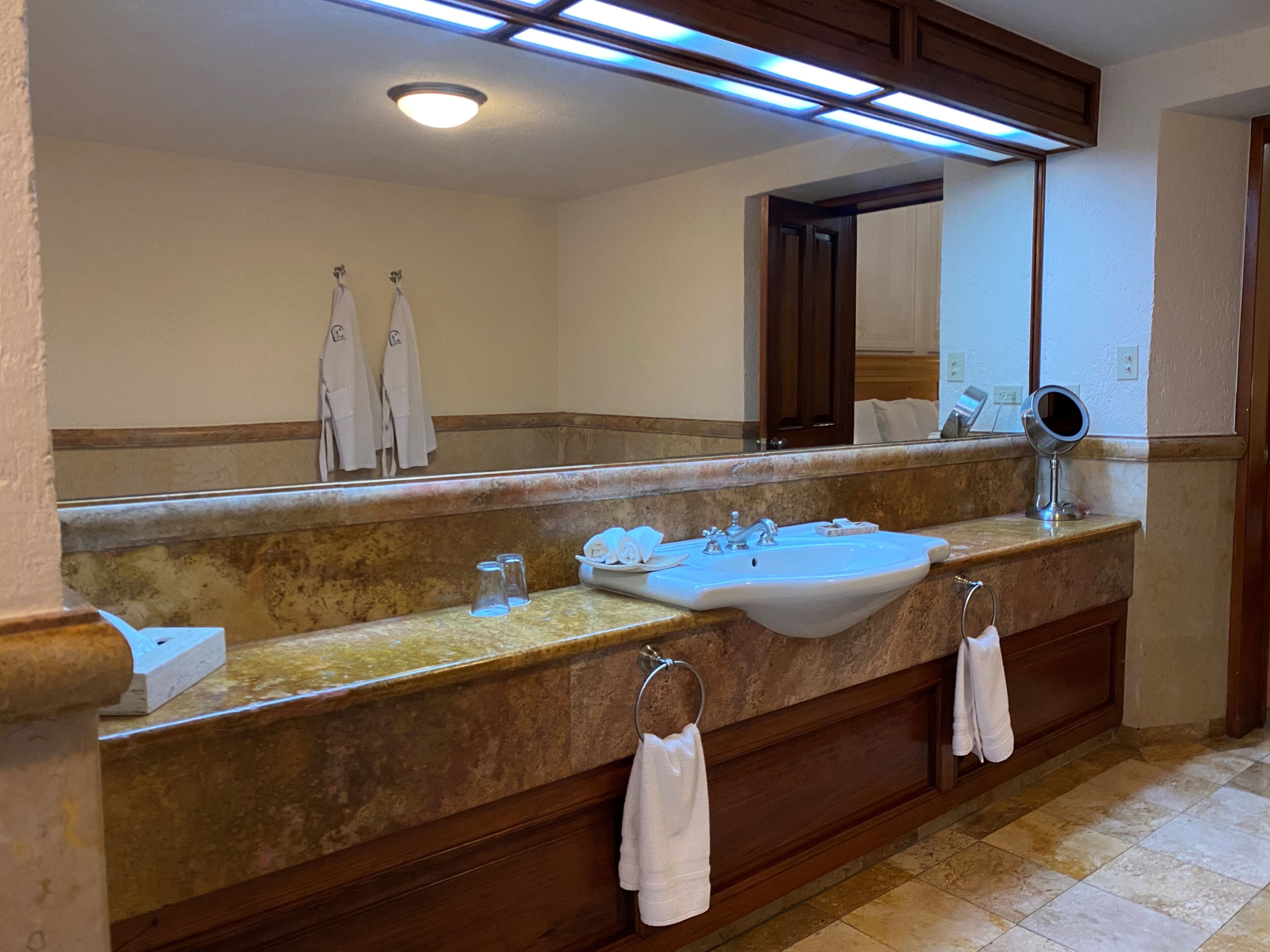 Un baño con un espejo grande, un lavamanos y tocador. Hay toallas colgadas en la pared y un perchero.