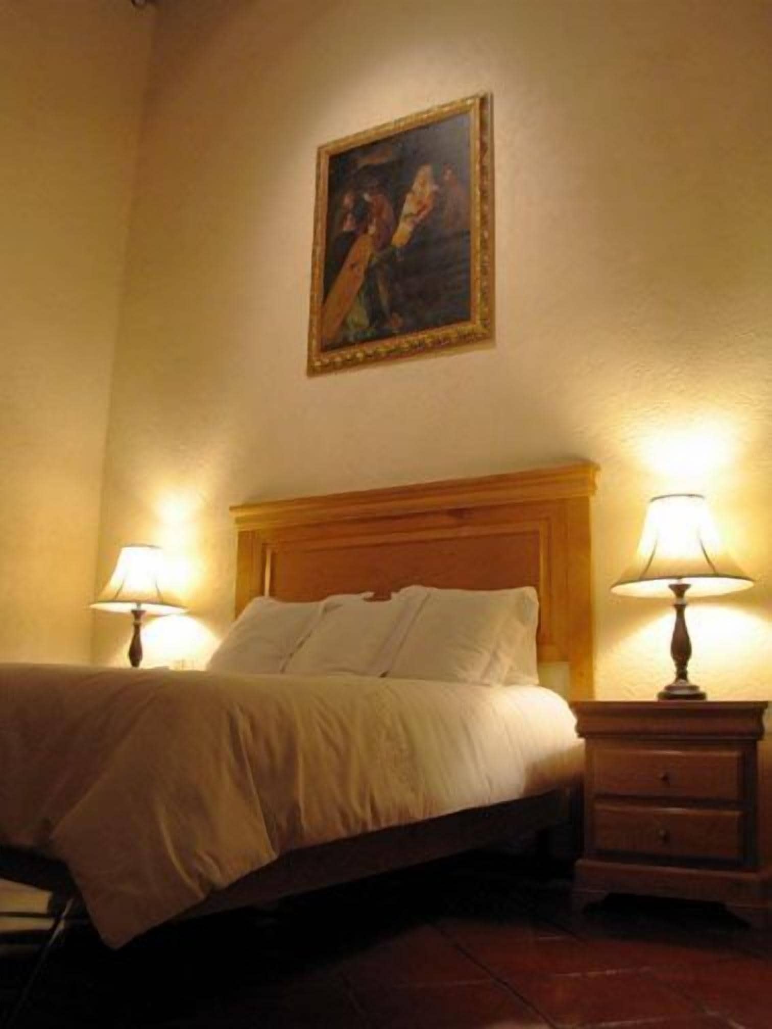 A hotel room with a bed, two bedside tables with lamps, a wooden headboard, and a painting on the wall.