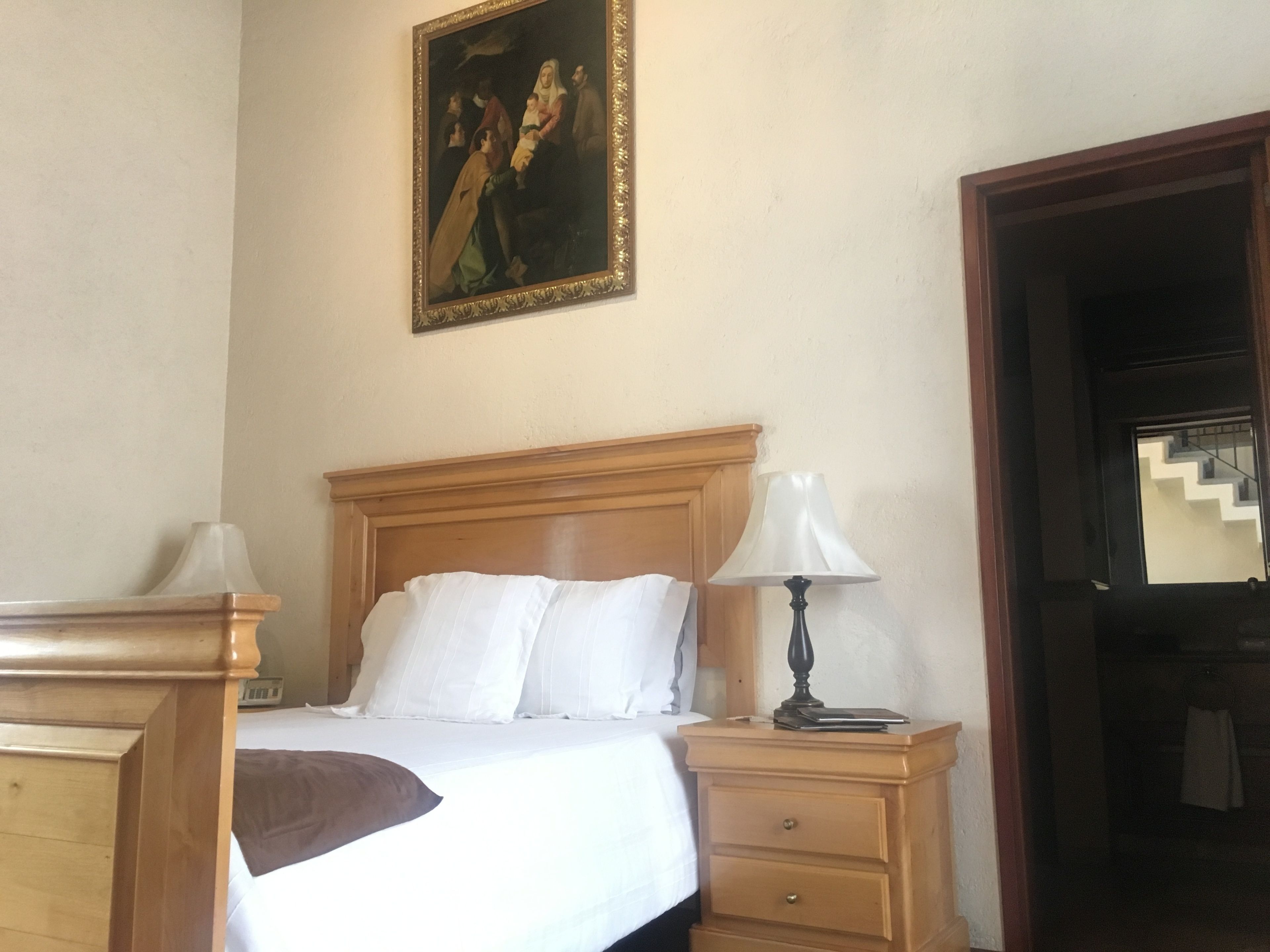 Habitación de hotel con cama de madera, ropa de cama blanca, mesita de noche con lámpara y un cuadro en la pared.